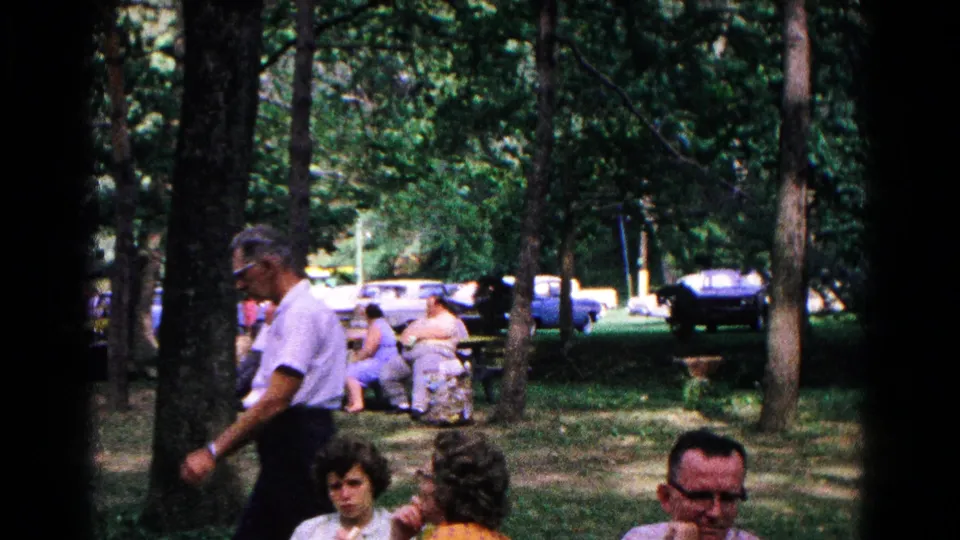 1963 Cold Springs home-movie frame showing adults seated in a shaded park as a man walks past trees with cars beyond.