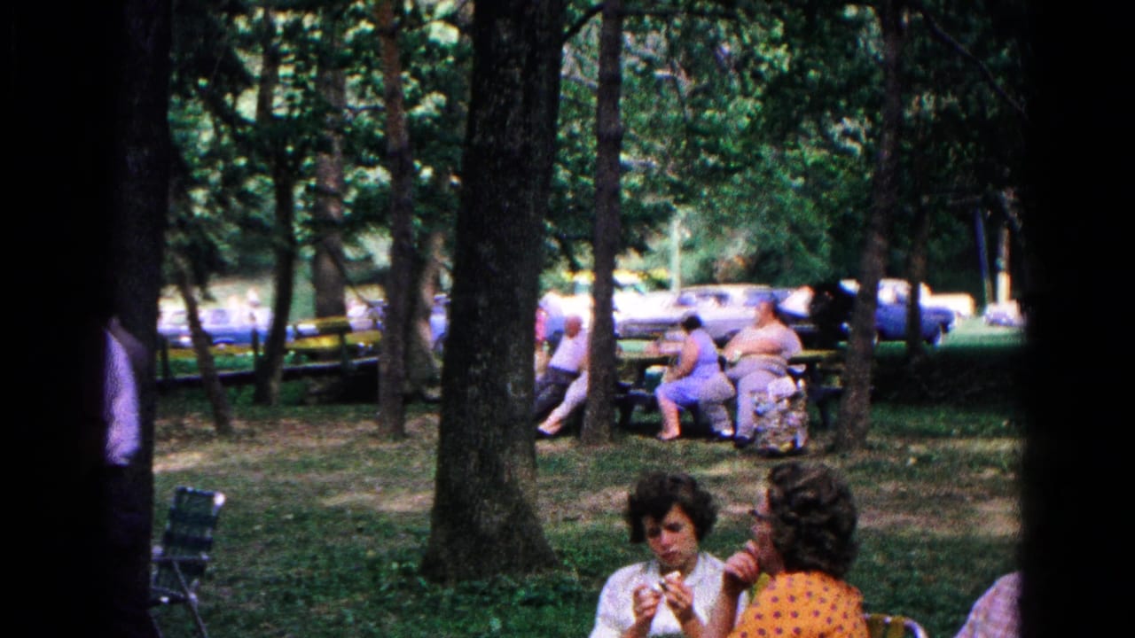 1963 Cold Springs frame showing women seated in folding chairs in a wooded park, with other adults and parked cars in the background.
