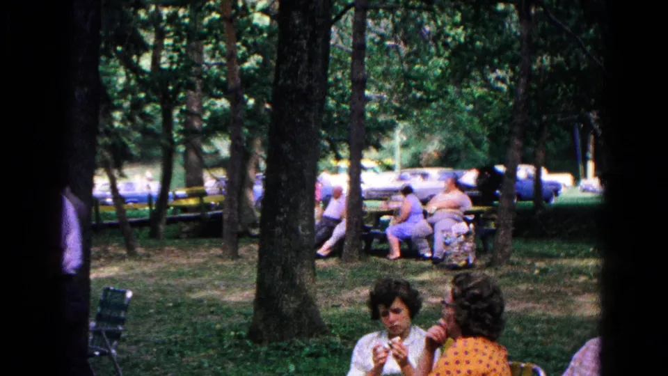 1963 Cold Springs frame showing women seated in folding chairs in a wooded park, with other adults and parked cars in the background.