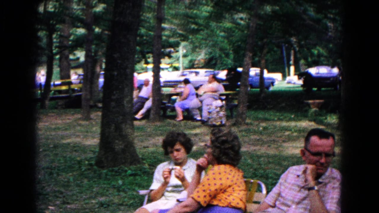 Adults seated in folding chairs at a wooded campground with parked cars behind them.
