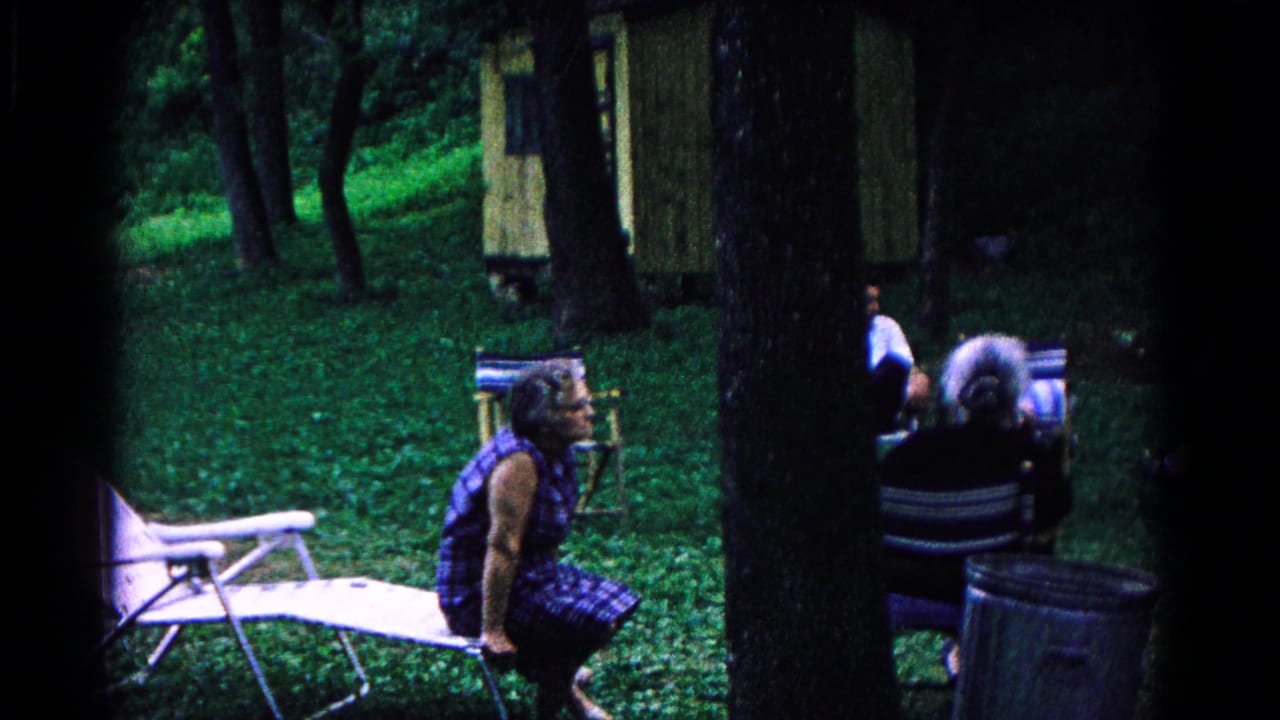 At the Cold Springs campground, seated adults appear near folding chairs, trees, a small structure, and a grassy shaded clearing.
