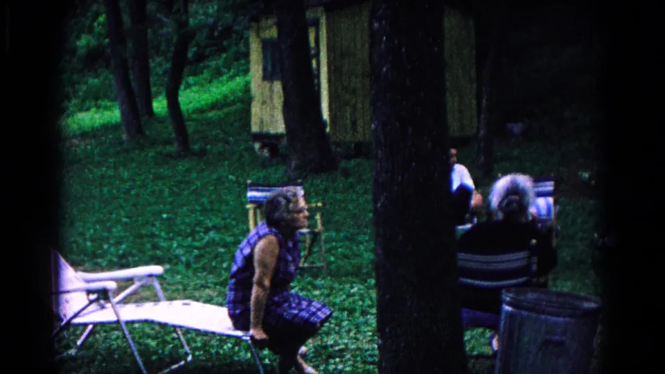At the Cold Springs campground, seated adults appear near folding chairs, trees, a small structure, and a grassy shaded clearing.