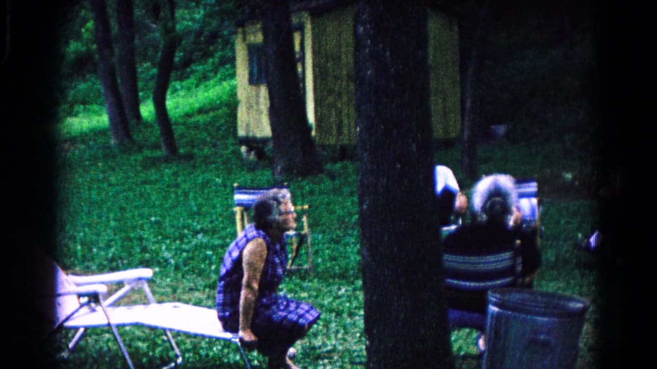A wooded 1963 Cold Springs campground view shows adults seated near portable chairs, trees, grass, and a small structure.