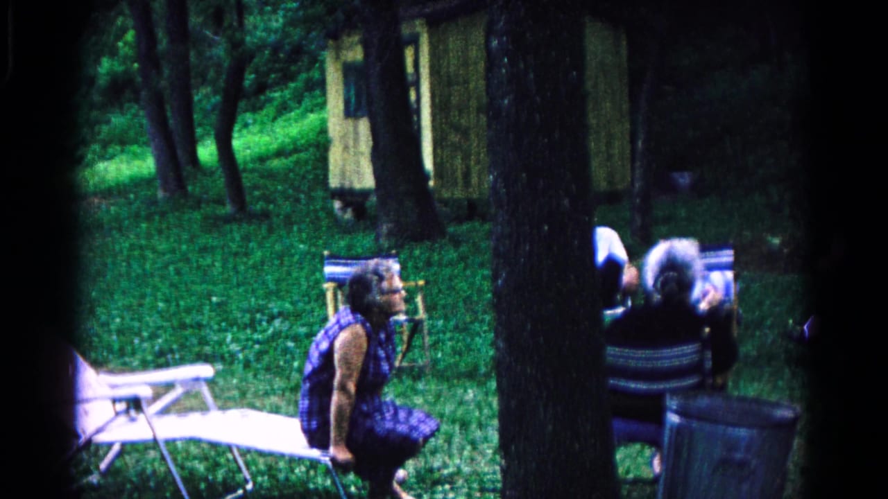 Older woman seated near folding chairs in a grassy wooded clearing.
