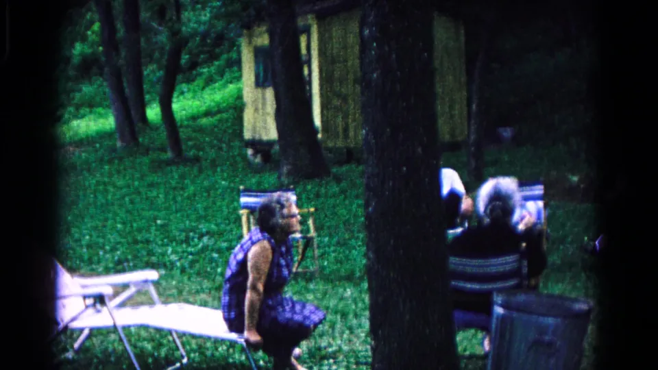 1963 Cold Springs home-movie frame of an older woman in a blue patterned dress seated near folding chairs under trees.