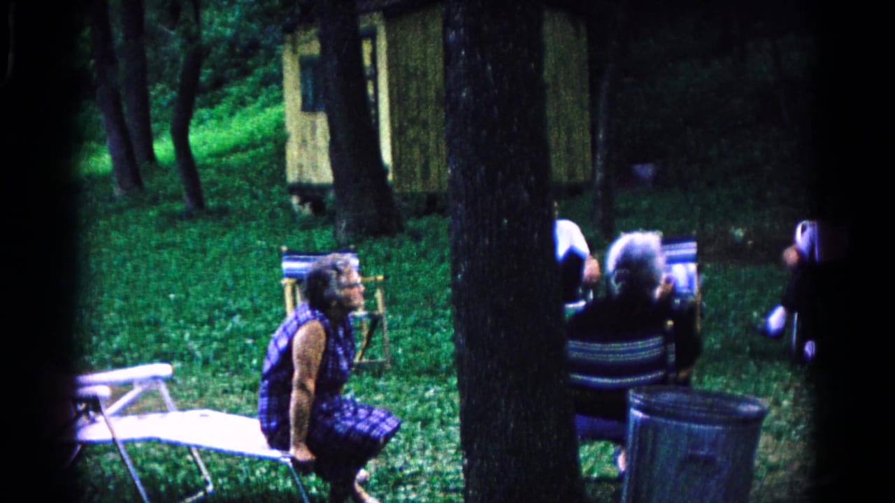 Adults seated in folding chairs near trees with a small cabin behind them.