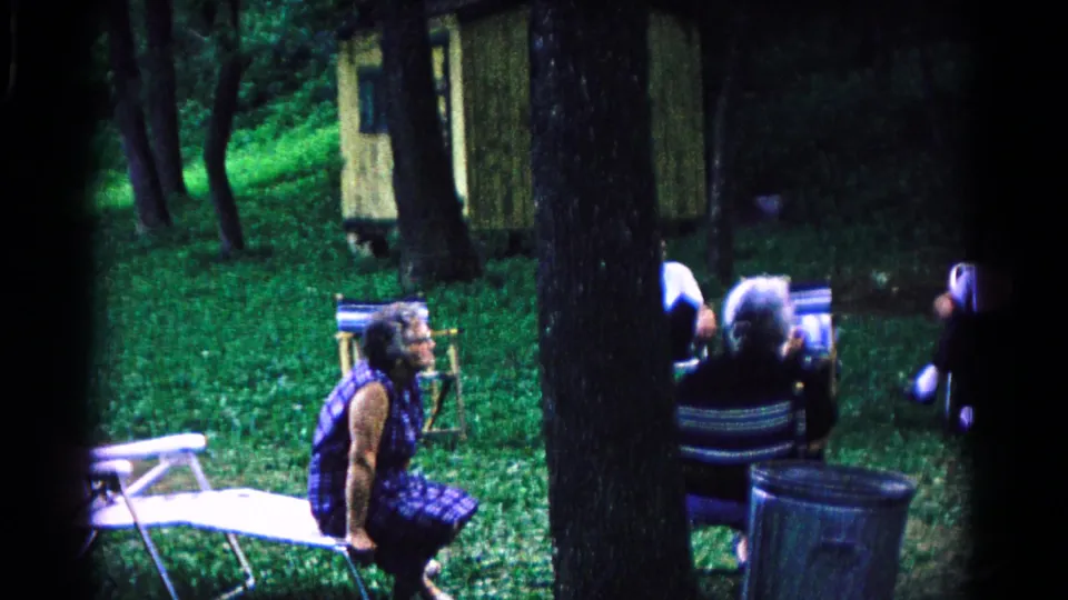 1963 Cold Springs frame showing older adults seated in folding chairs in a shaded grassy clearing with a small cabin in back.