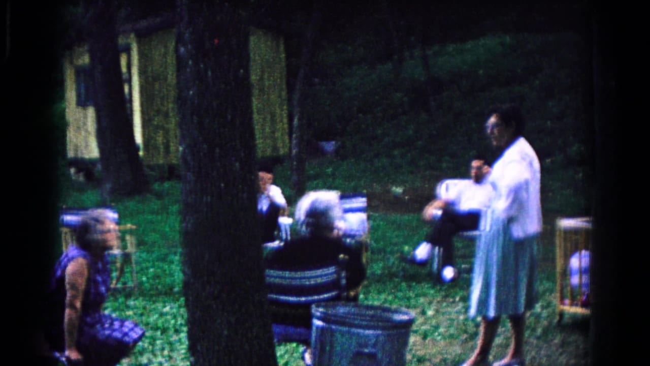 1963 Cold Springs home-movie frame with older adults in lawn chairs and a man standing in a light coat among trees.