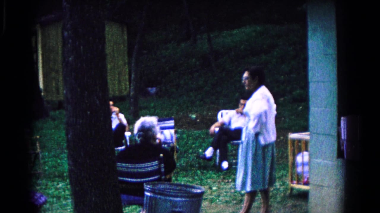 1963 Cold Springs frame of a man standing beside seated adults under trees, with outdoor chairs and a building edge visible.