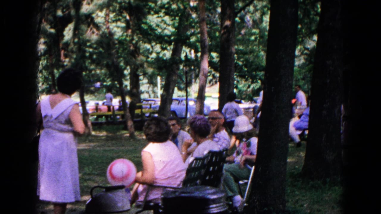 1963 Cold Springs park scene with people seated and standing under trees, a striped balloon, benches, and parked cars in the background.