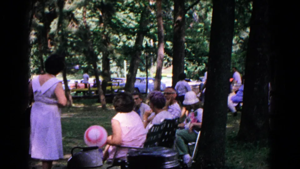 1963 Cold Springs park scene with people seated and standing under trees, a striped balloon, benches, and parked cars in the background.