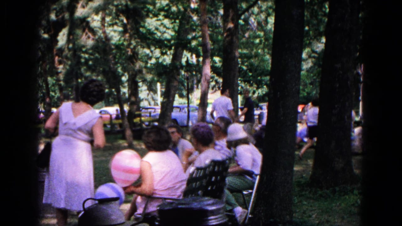 1963 Cold Springs home-movie frame showing a striped balloon, seated adults, benches, trees, and cars beyond the picnic area.