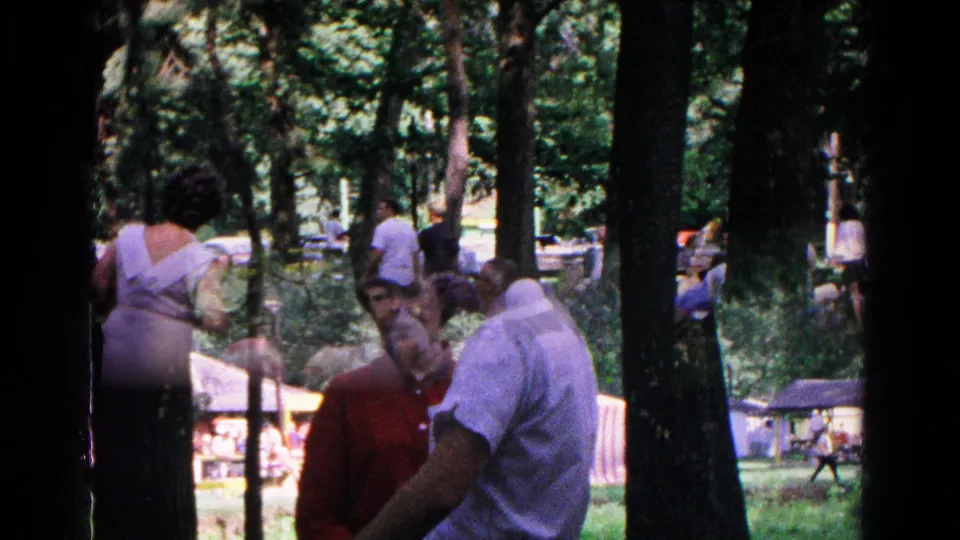 1963 Cold Springs park gathering with adults in short-sleeve clothing, benches, tall trees, and a light structure in the background.