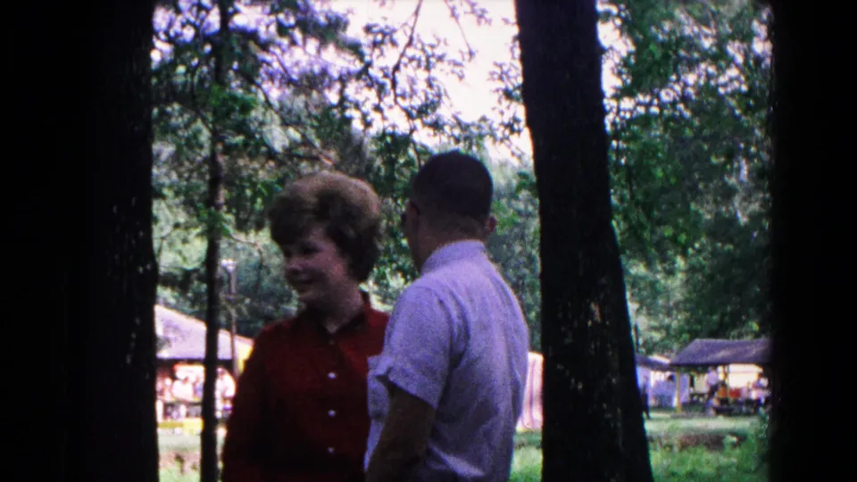 1963 Cold Springs home-movie frame with a woman in red and a man in a light shirt standing near trees and park structures.
