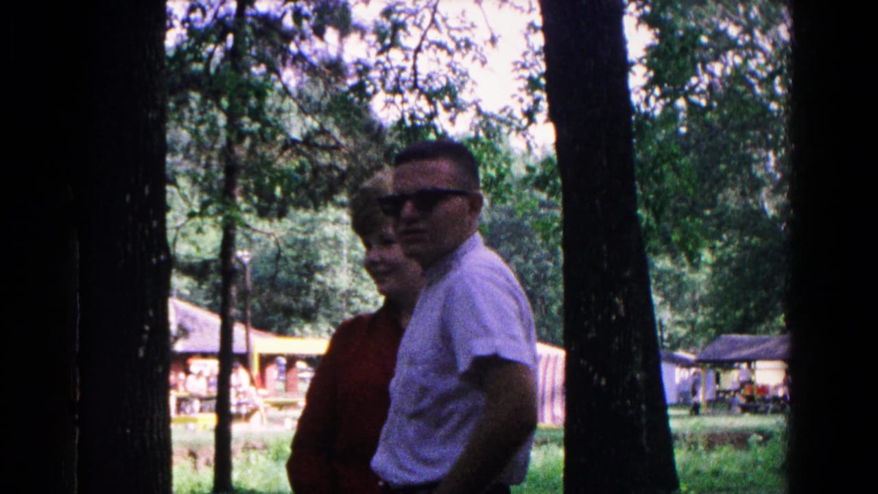 A woman in red and a man in sunglasses stand close together among trees in a park.