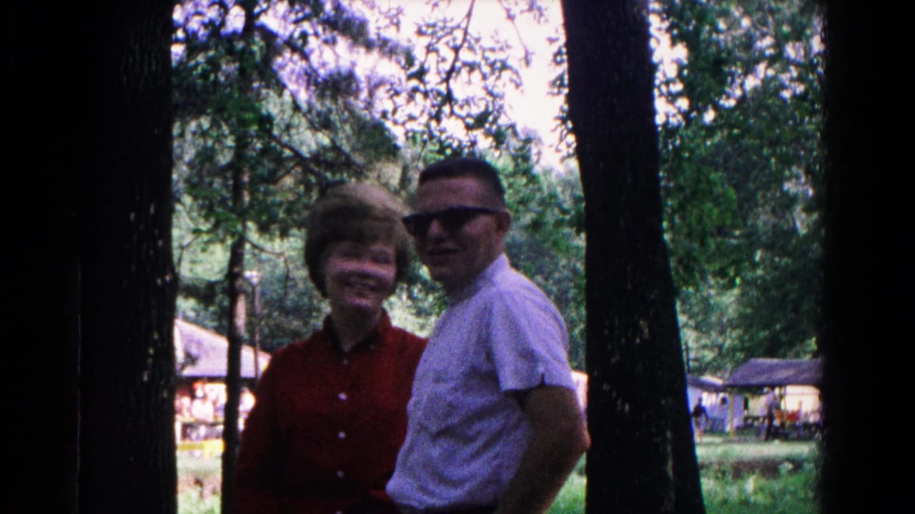 The couple faces the camera and smiles in a wooded outdoor setting.