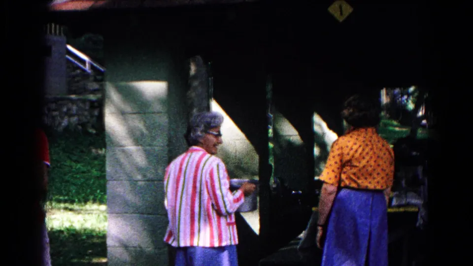 The women remain near the covered food-service window in Cold Springs, New York, with bright grass and stonework visible at the edge of shade.