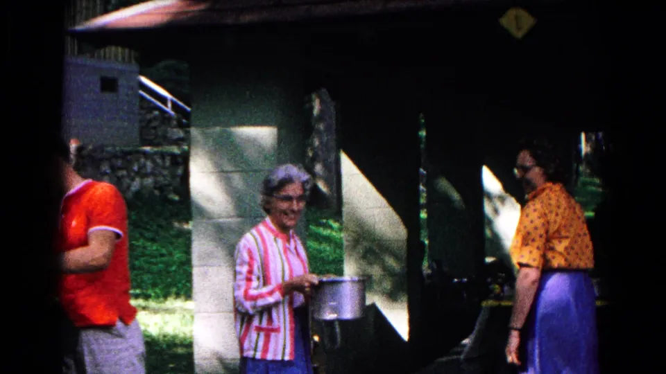 A woman in a striped top smiles or turns toward the camera while holding a pot, with another woman and a partial passerby at the shaded stand.
