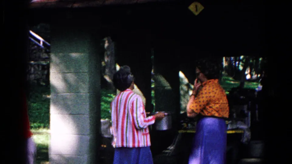 At the covered outdoor food stand, the striped-shirt woman holds a metal pot while the woman in yellow stands beside the dark opening.
