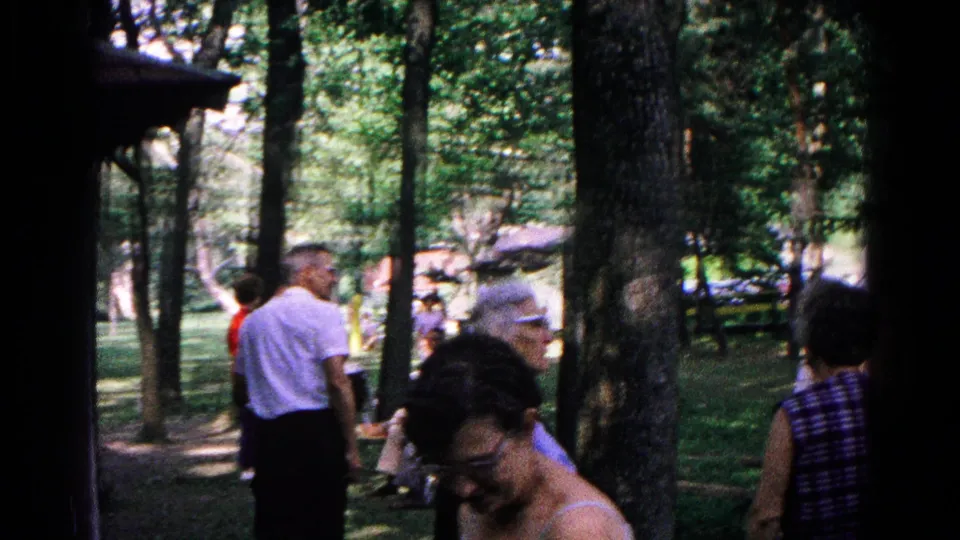 Adults move through a shaded wooded area, with a partial masonry structure at left and benches deeper in the Cold Springs park scene.