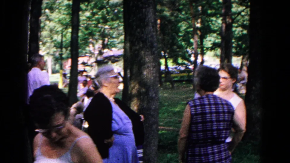 Women in sleeveless dresses and a man in a white shirt stand in a wooded Cold Springs park during the 1963 home-movie gathering.