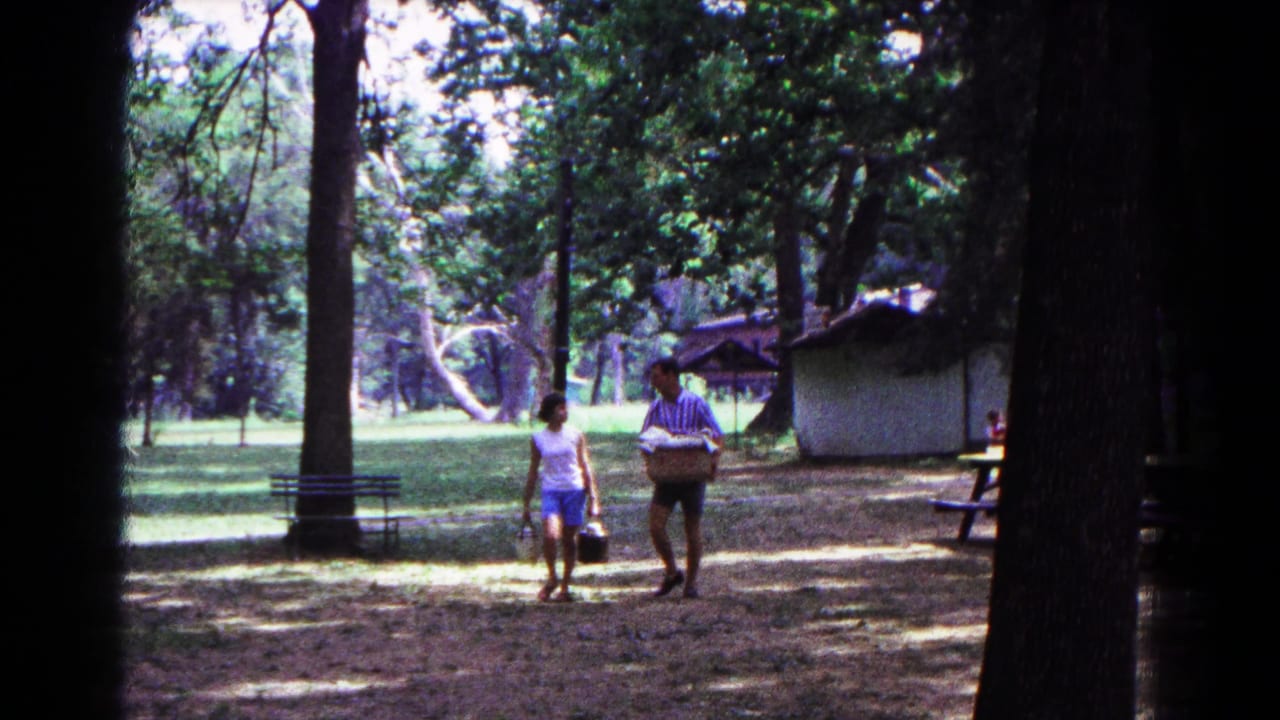 Two adults walk through a wooded campsite while one carries a picnic basket.