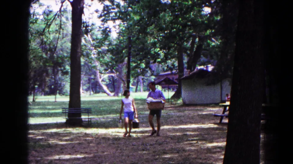 1963 Cold Springs home-movie frame of two adults walking under tall trees near benches and a small shelter at a wooded campsite.