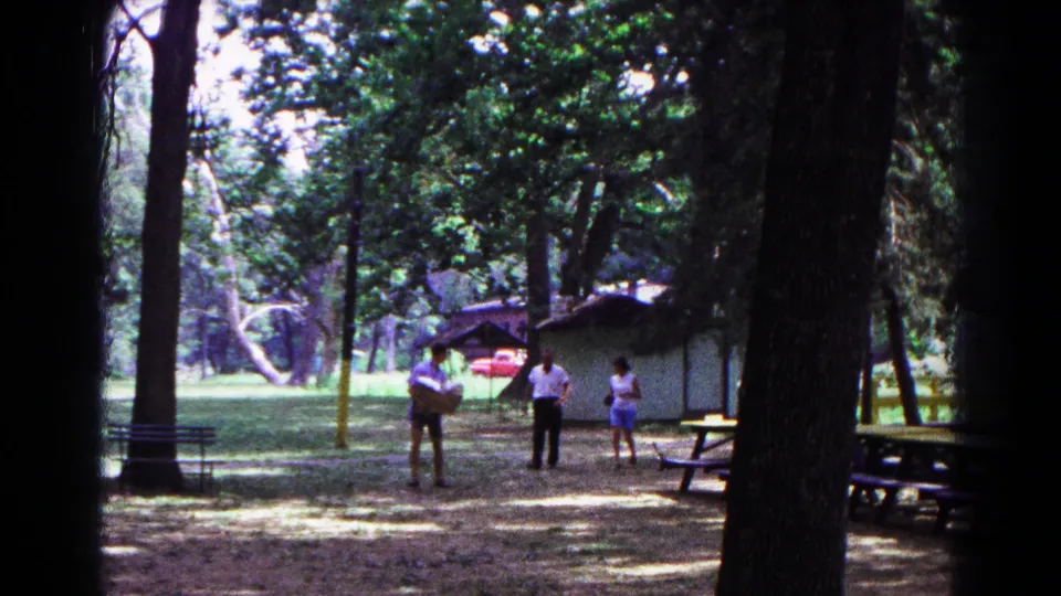 1963 Cold Springs home-movie frame with adults near a small shelter, picnic tables, tall trees, and one person holding a basket.
