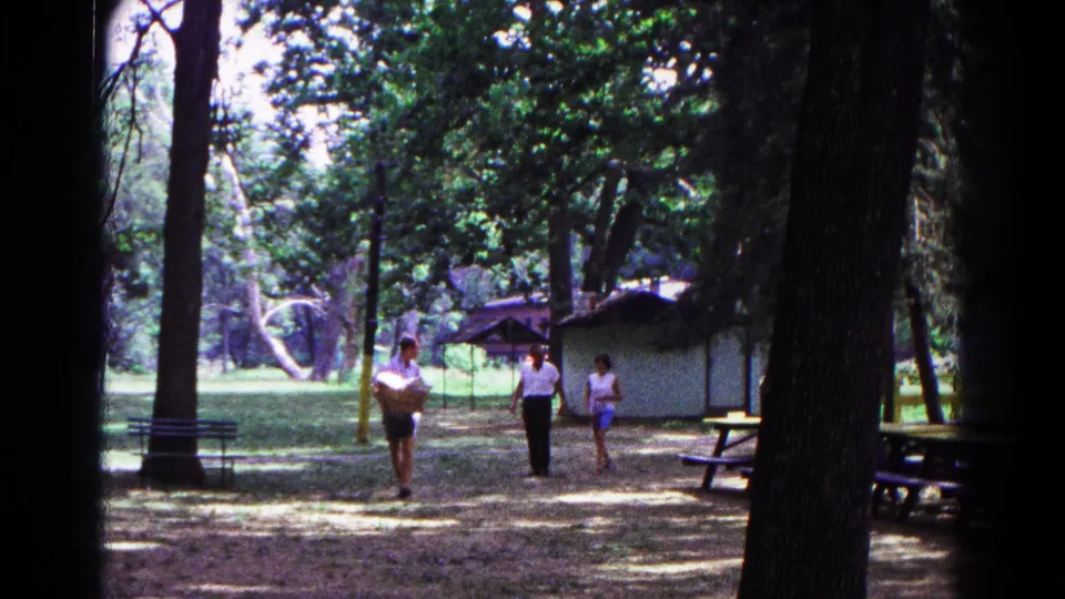 Final 1963 Cold Springs frame of three adults walking through a campsite, with tall trees, tables, and a small shelter in the background.
