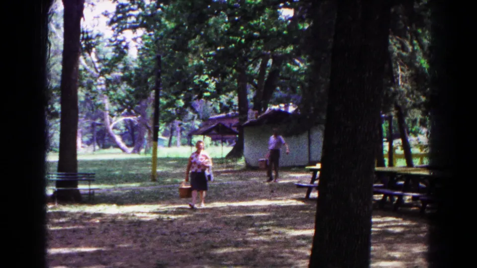 1963 Cold Springs, New York home-movie frame showing two adults approaching through a wooded picnic area with a basket and park shelter.