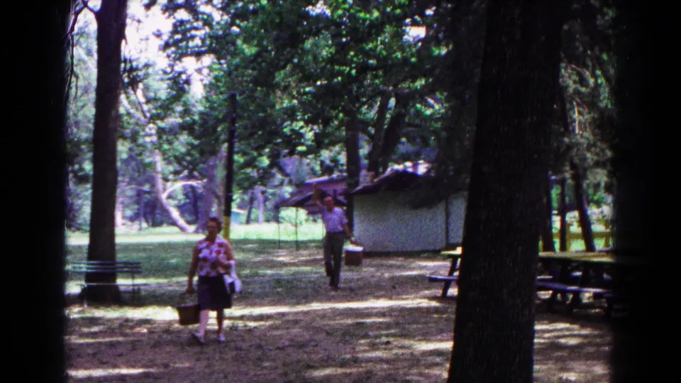 1963 Cold Springs, New York frame showing adults nearing the camera in a shaded park picnic area with tables and a shelter behind them.