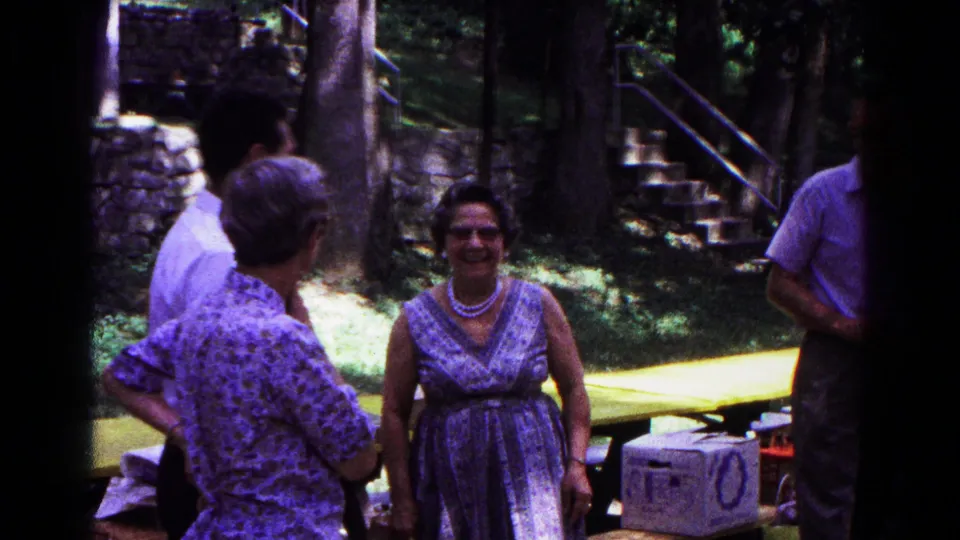 1963 home-movie frame from Cold Springs, New York, showing adults talking near a yellow picnic table under trees.