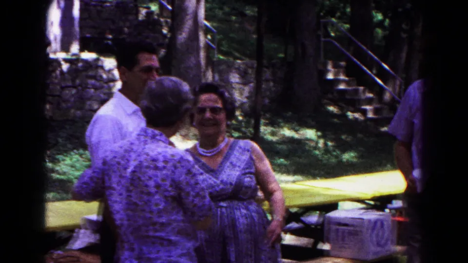 1963 Cold Springs park gathering with a smiling woman in a patterned dress and pearl necklace near adults and a cooler.