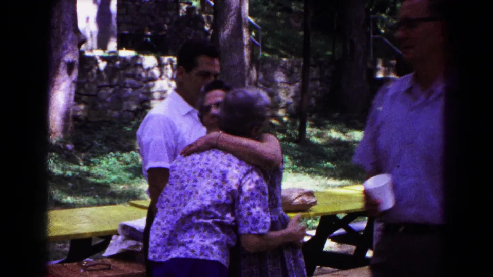 Cold Springs 1963 home-movie frame showing adults greeting each other near a yellow picnic table in a shaded park.