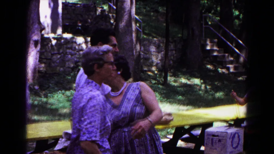 1963 Cold Springs park scene with adults embracing beside a yellow picnic table, trees, stonework, and a white cooler.
