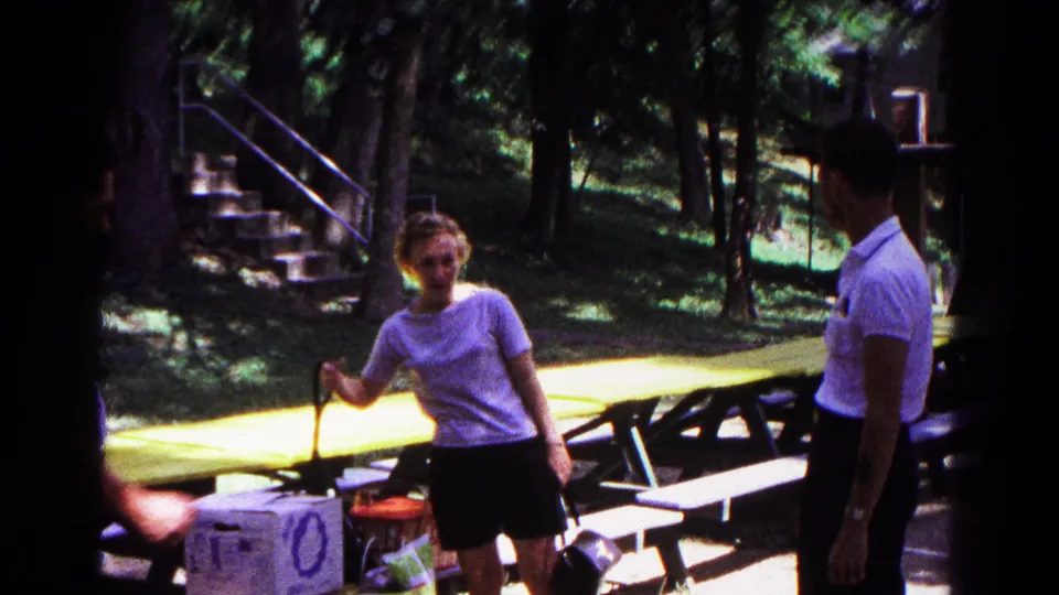 1963 8mm frame from Cold Springs showing adults near yellow picnic tables, trees, steps, and a cooler in dappled sunlight.