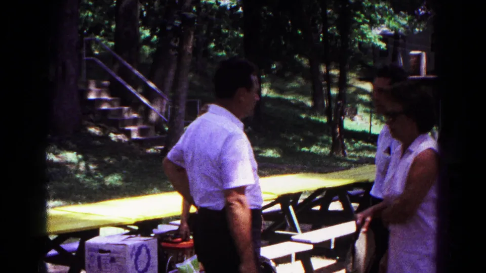 A man in a white short-sleeved shirt and a woman in sunglasses stand near yellow picnic tables in the wooded Cold Springs setting.