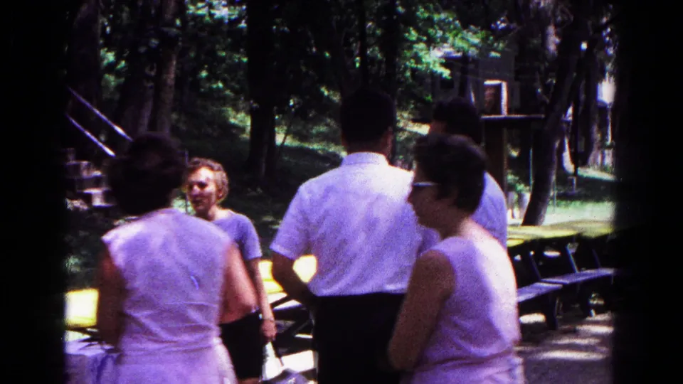 Several adults in 1960s casual clothing stand around picnic tables, with trees and stone steps visible behind them.