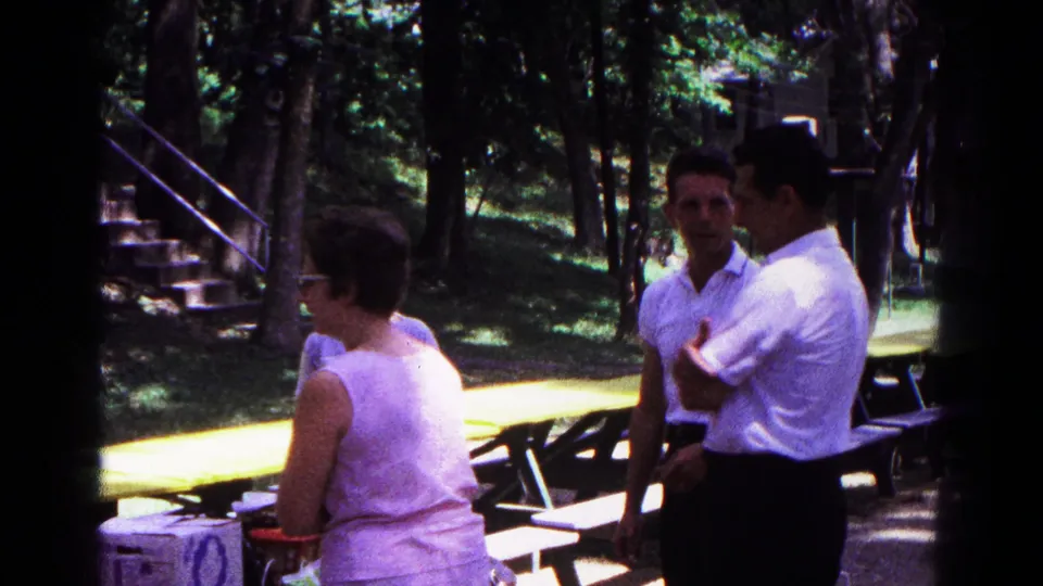 Adults stand near yellow picnic tables in Cold Springs, with a wooded background and stone steps visible in the home-movie frame.