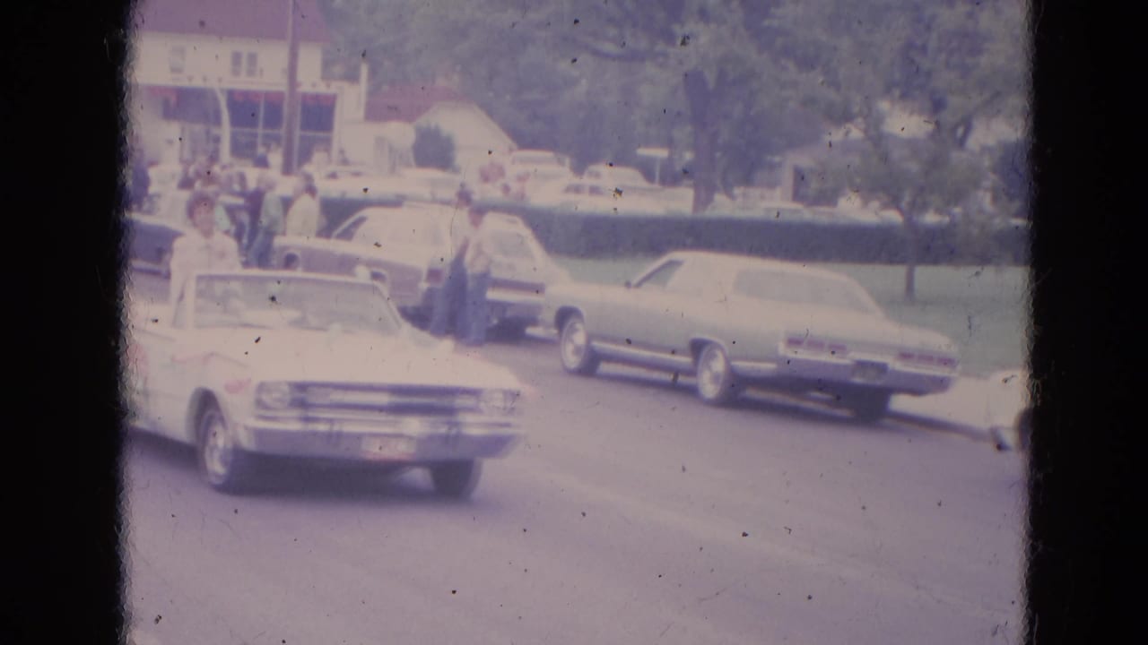 White convertible and parked cars on a parade route with spectators in the background.
