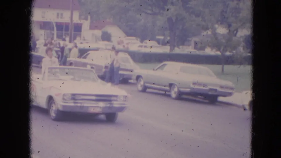 1963 Lincoln, Nebraska parade view with a white convertible entering frame, parked cars, spectators, trees, and a distant building.