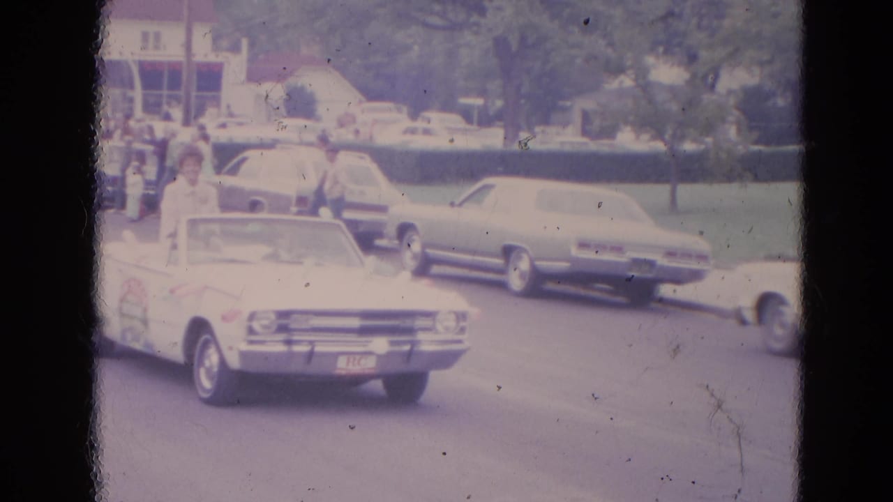 White convertible passing parked mid-century cars on a street with spectators nearby.