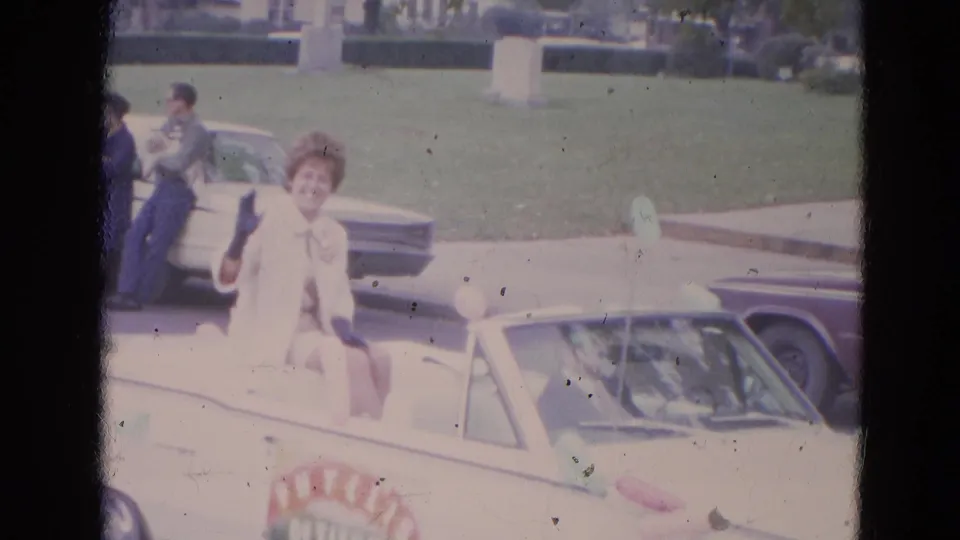 A woman in formal light-colored clothing and gloves waves from the back of a white convertible during the 1963 Lincoln, Nebraska parade.