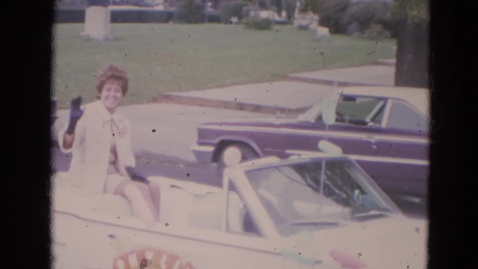 The woman continues waving from the convertible as the car passes a curb, lawn, sidewalk, tree, and parked mid-century automobile.