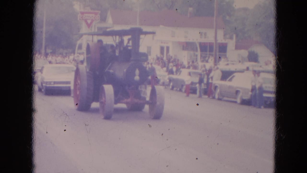 Steam tractor approaches on a parade street with spectators, parked cars, and a Conoco sign behind it.