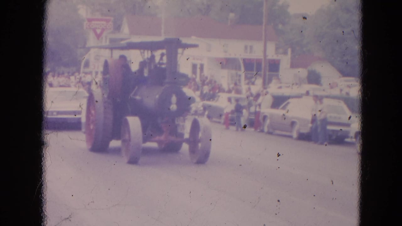 Steam tractor continues down the street, with people on sidewalks and classic cars parked along the curb.