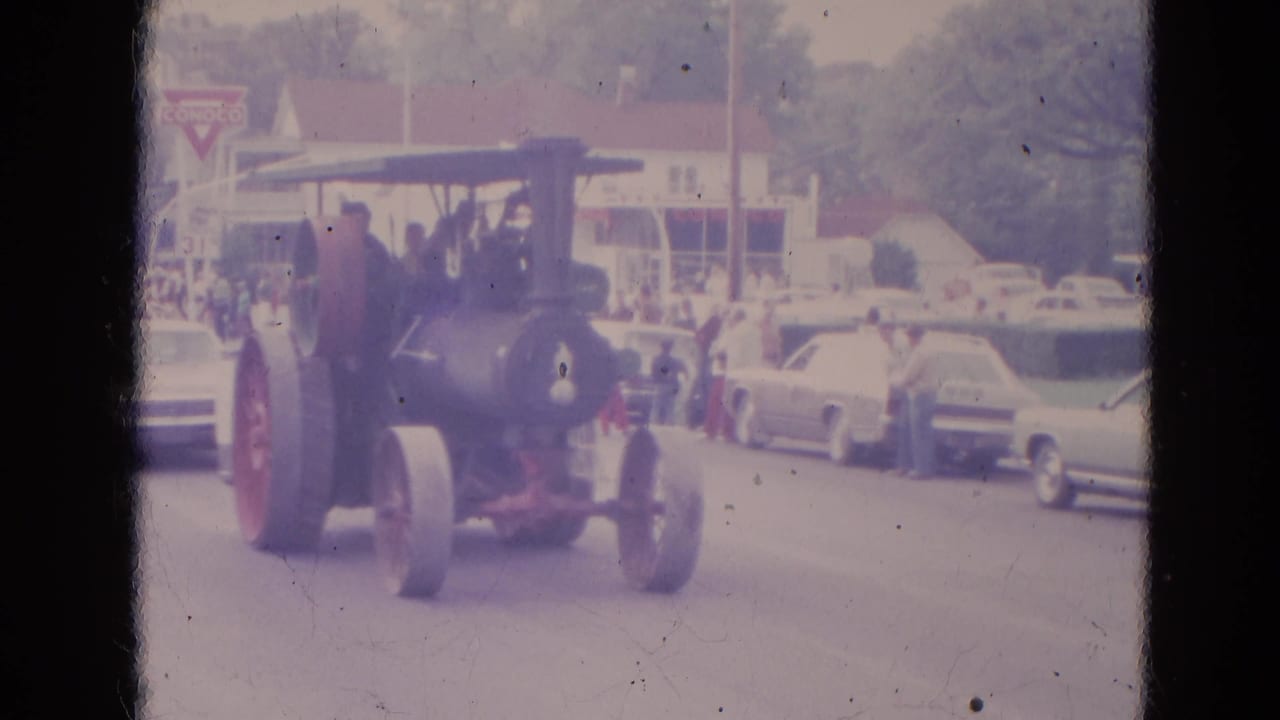 Steam tractor with large red wheels driving along a parade route past parked cars and spectators.