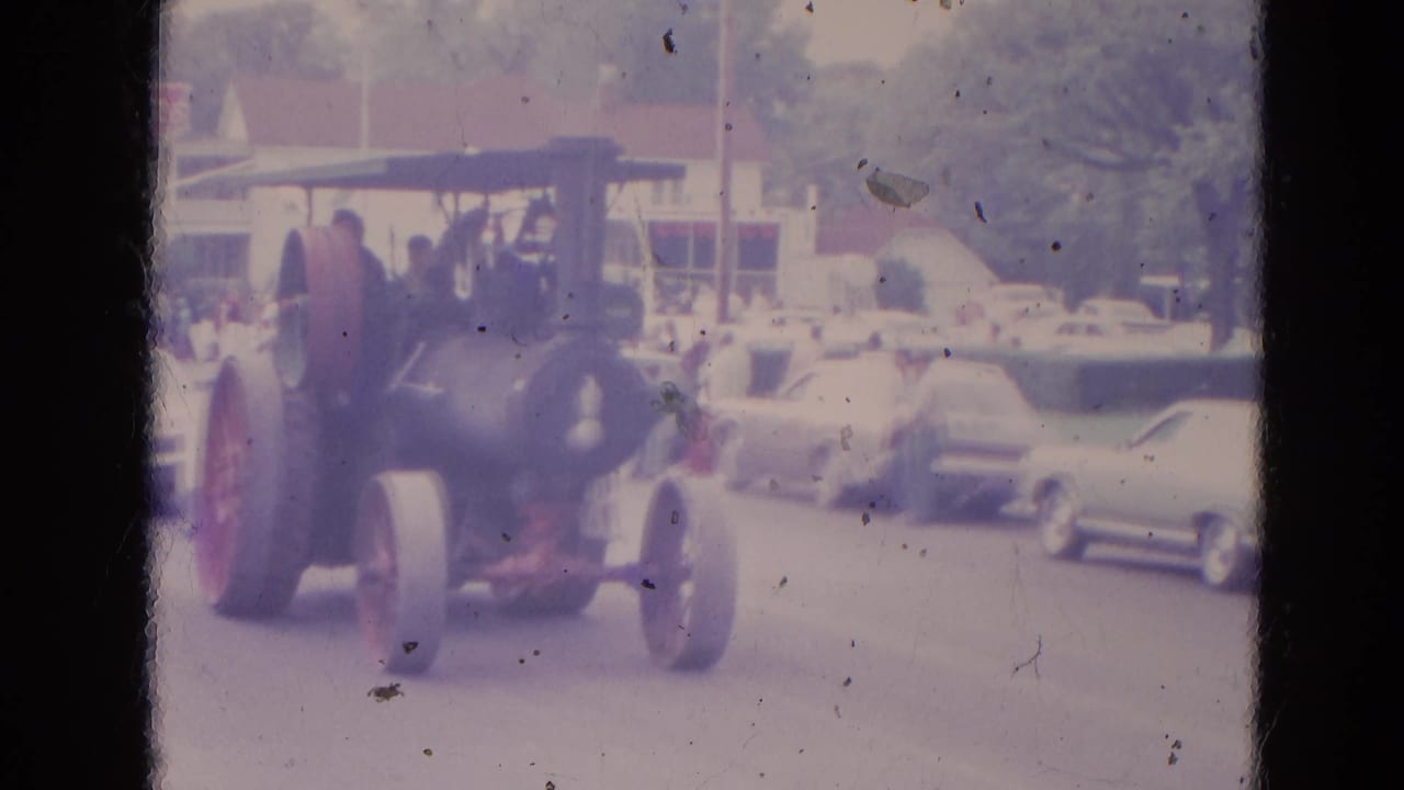 Steam tractor continues along the street with parked cars and spectators in the background.