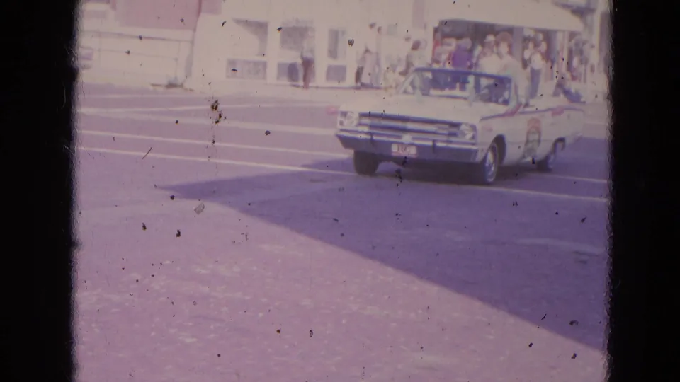 A white convertible with parade passengers moves through a Lincoln, Nebraska street scene in this 1963 8mm home-movie frame.