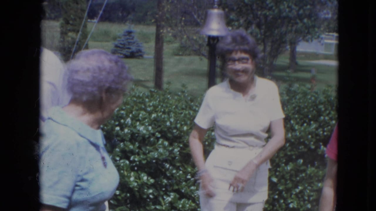 Adults stand in a garden near hedges, with a woman in white facing the camera.